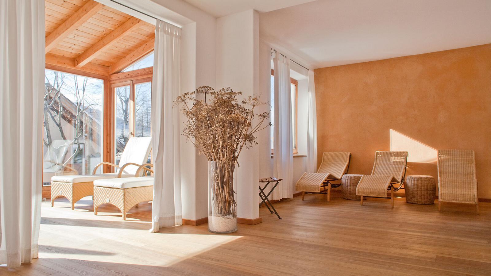 Wellness area with wicker beds, large bright windows, white curtains and potted vase with dry flowers in our 3-star wellness hotel in Sesto Pusteria