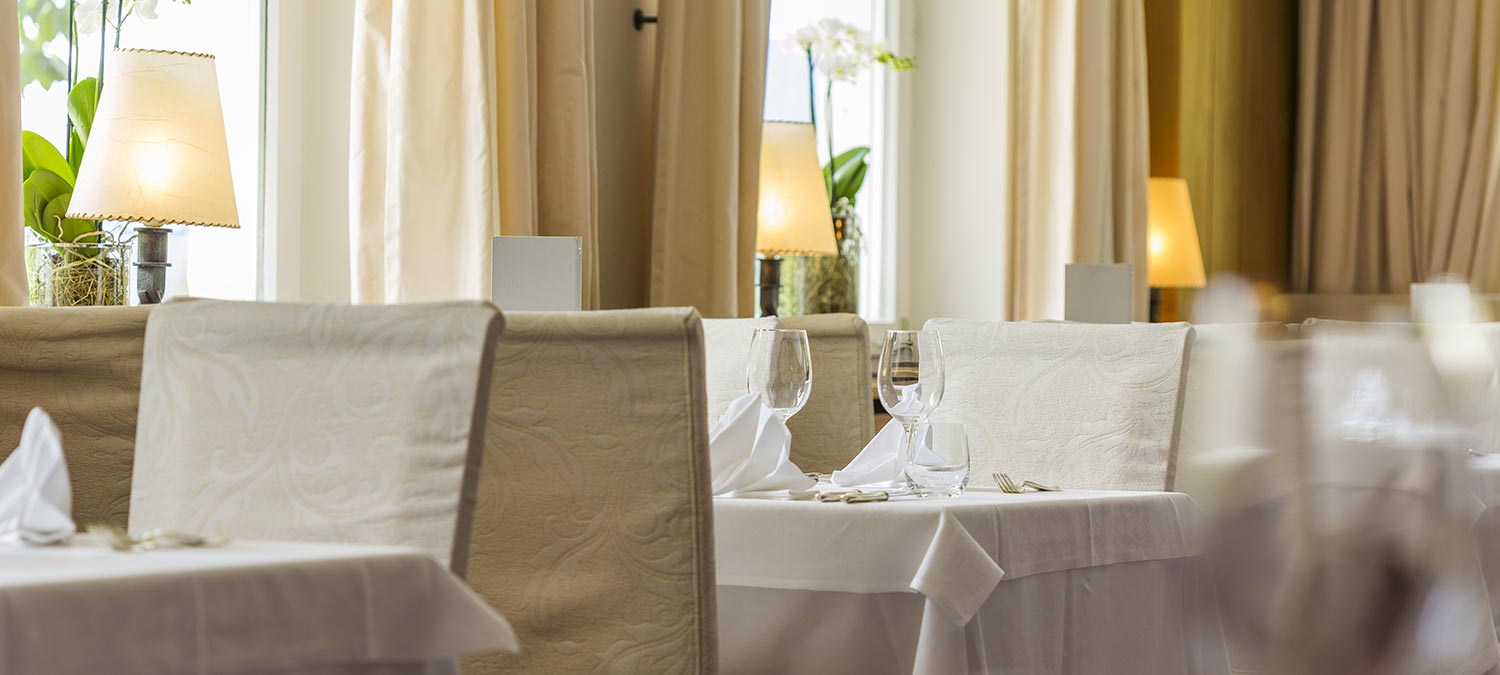 Detail of a table in our hotel's dining room in Sesto Pusteria with white tablecloth, cream-coloured chairs and matching curtains