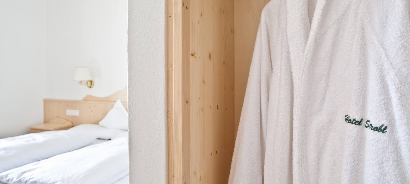 Detail of a white cotton bathrobe hanging in one room of our holiday apartments in Sesto Pusteria furnished in spruce with double bed in the background