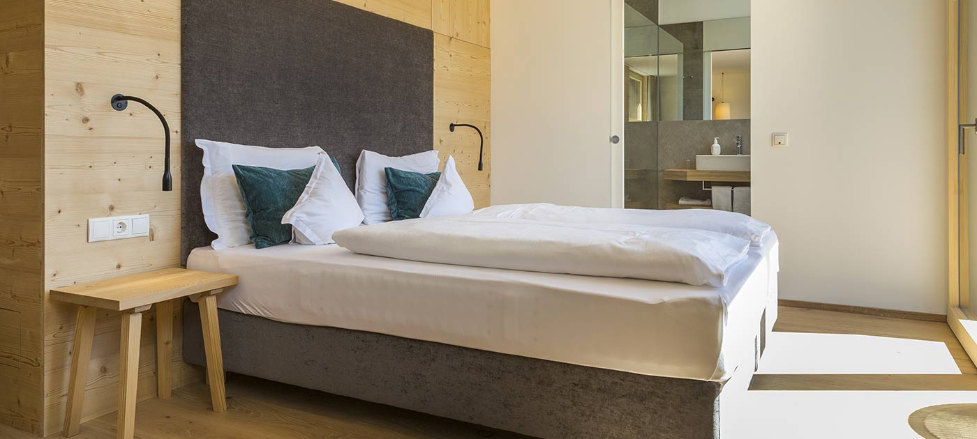 Gemütliches und geräumiges Zimmer im 3-Sterne-Hotel Strobl im Sexten im Pustertal mit Holzmöbeln, geräumigem Doppelbett und elegantem Badezimmer im Hintergrund