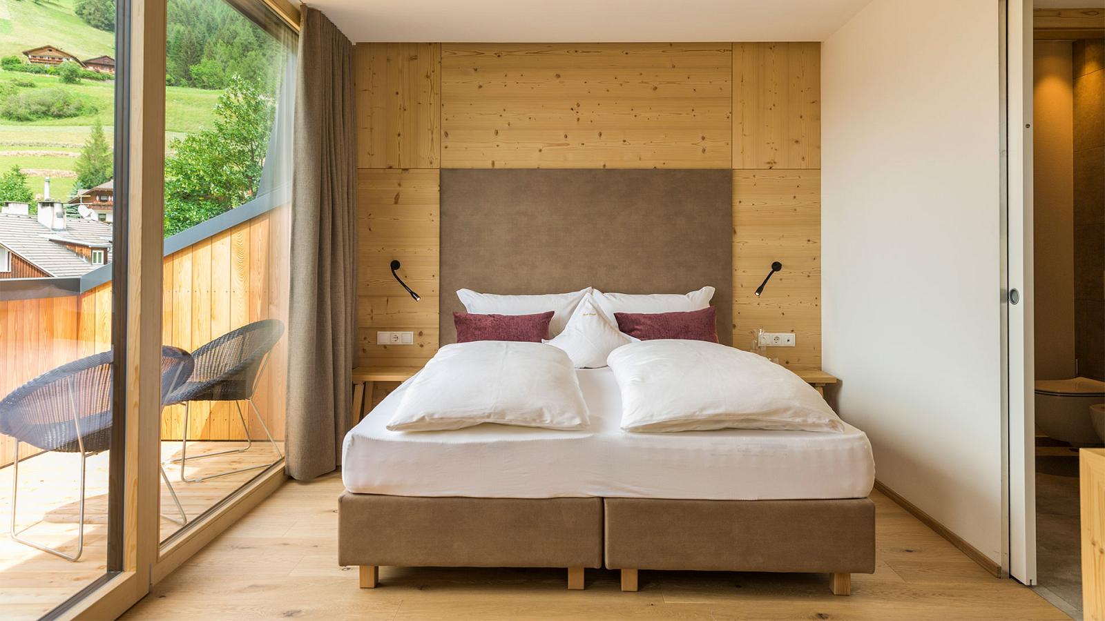 Una confortevole camera dei nostri appartamenti a Sesto in Val Pusteria con letto matrimoniale, parete di legno e accesso con porta finestra al comodo balcone soleggiato