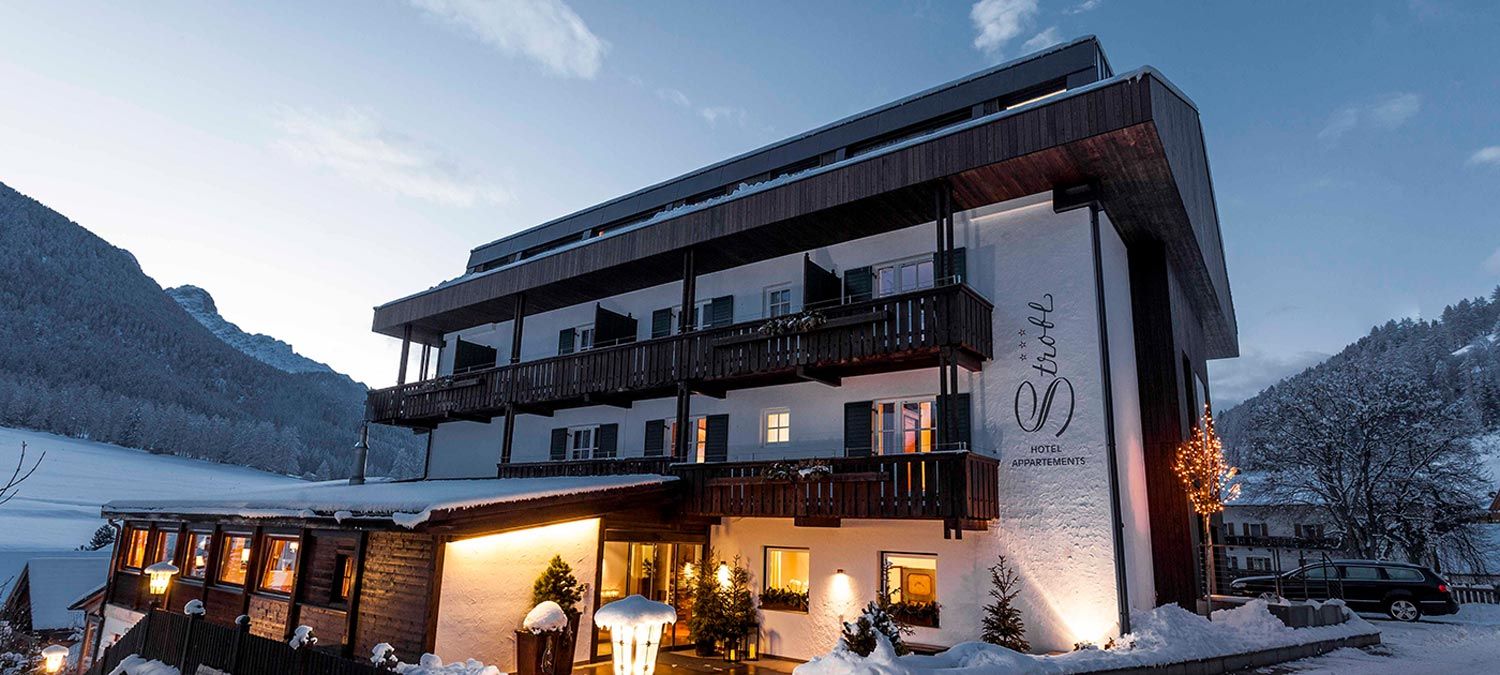 Fassade des Hotels Strobl in Sexten im Winter, mit beleuchteten Fenstern und im Hintergrund Wiesen und schneebedeckte Wälder
