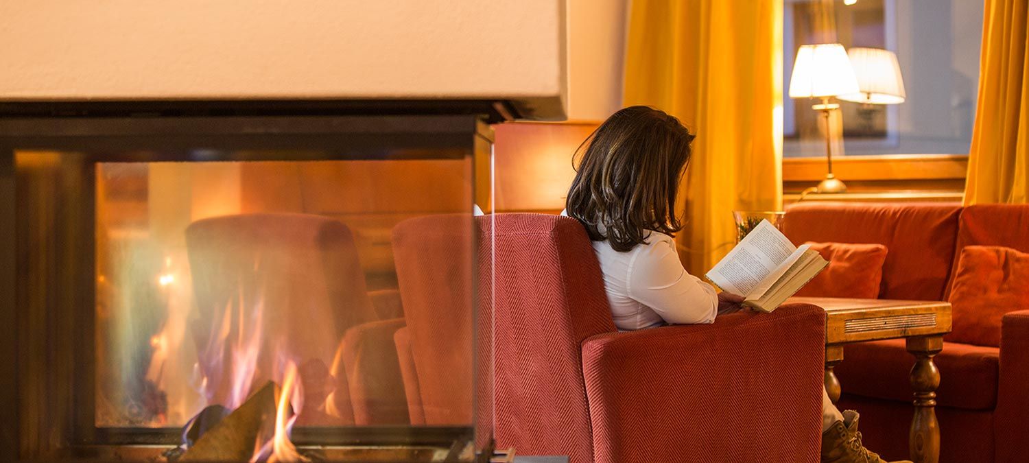 Eine Frau liest ein Buch vor dem Kamin auf dem weichen roten Sessel unserer traditionellen Südtiroler Stube